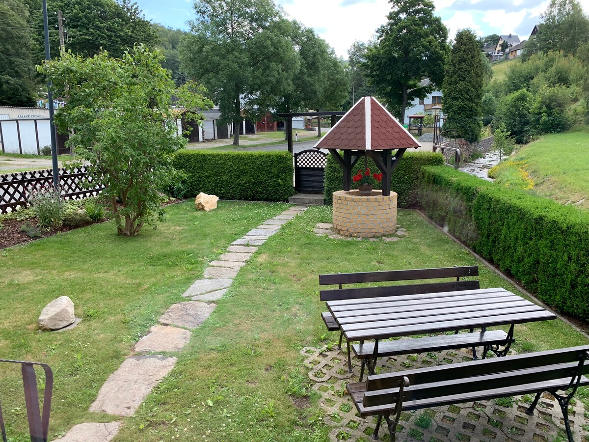 A well-maintained garden features a stone pathway leading to a covered well. Wooden benches surround a patio area, surrounded by neatly trimmed hedges and trees. The grass is lush, providing a serene outdoor space, with glimpses of surrounding nature in the background.