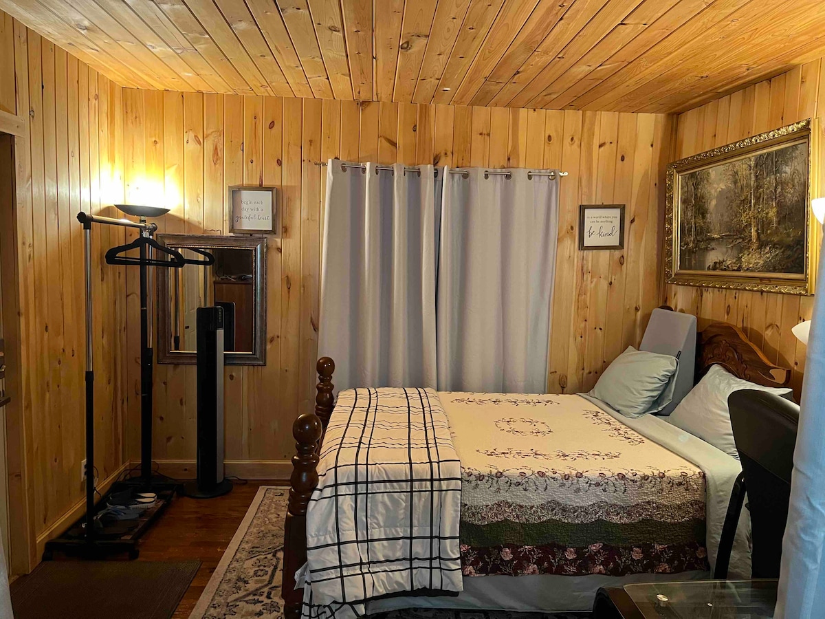 A cozy sleeping area is presented with knotty pine paneling and soft lighting. A spacious bed is adorned with a patterned blanket, complemented by light blue pillows. A clothing rack is positioned nearby, and curtains frame a window, allowing for privacy.