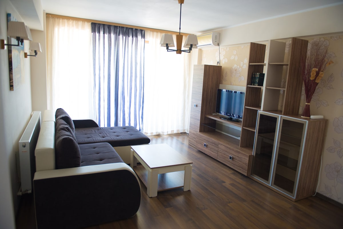 A living area features a dark sectional sofa accompanied by a light coffee table. A wall unit with shelves and a television creates a functional space. Floor-to-ceiling curtains in light colors allow natural light to fill the room. Wooden flooring adds warmth to the atmosphere.