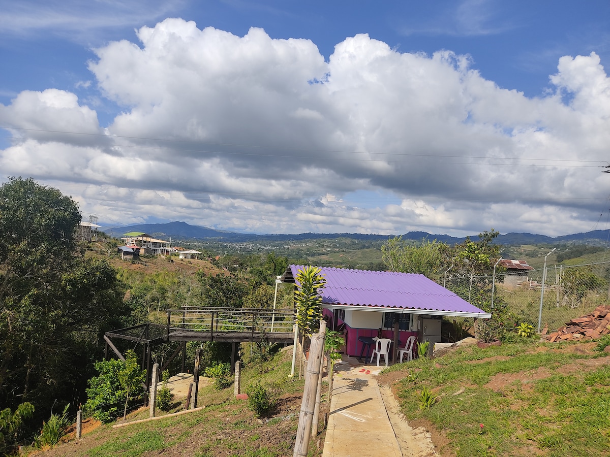 A small, independent structure is visible against a backdrop of rolling hills and scattered clouds. Surrounding greenery adds to the serene environment. A pathway leads from the foreground to the building, which features a purple roof and a porch with white chairs.