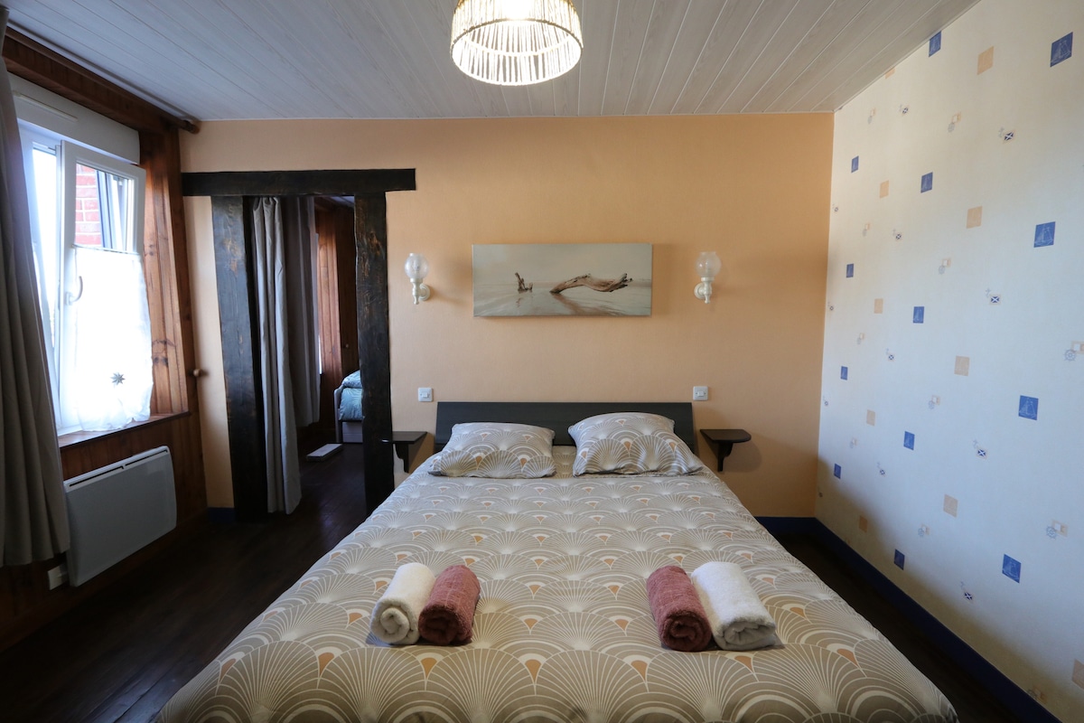 A spacious bedroom features a large bed with patterned linens, flanked by bedside sconces. Natural light enters through the window, revealing light-colored walls with decorative elements. A doorway leads to an adjoining room, and decorative towels are neatly arranged on the bed.