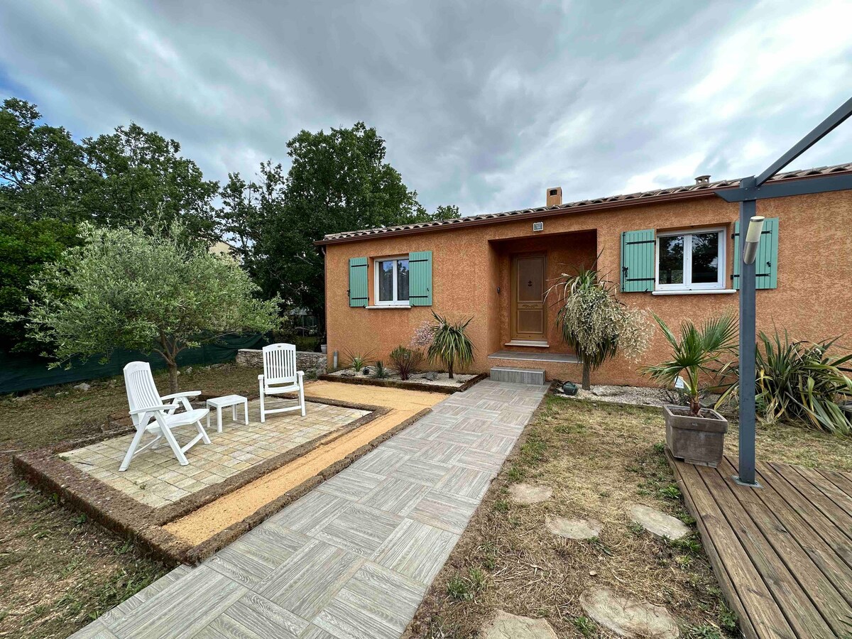 The exterior of a single-story home is featured, showcasing a welcoming entrance with a wooden door and surrounding greenery. A light-colored stone pathway leads to the door, flanked by two white lounge chairs. Vibrant plants are positioned near the entrance, creating a serene outdoor space.