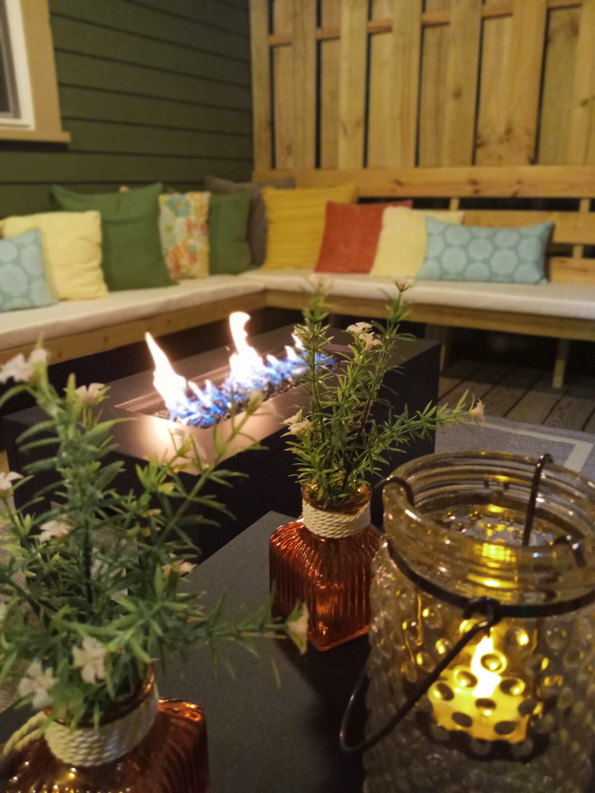 A cozy outdoor seating area is illuminated by the warm glow of a fire pit. A mix of colorful cushions adorns the bench, while small, elegant vases with greenery and a softly glowing lantern add ambiance to the space.