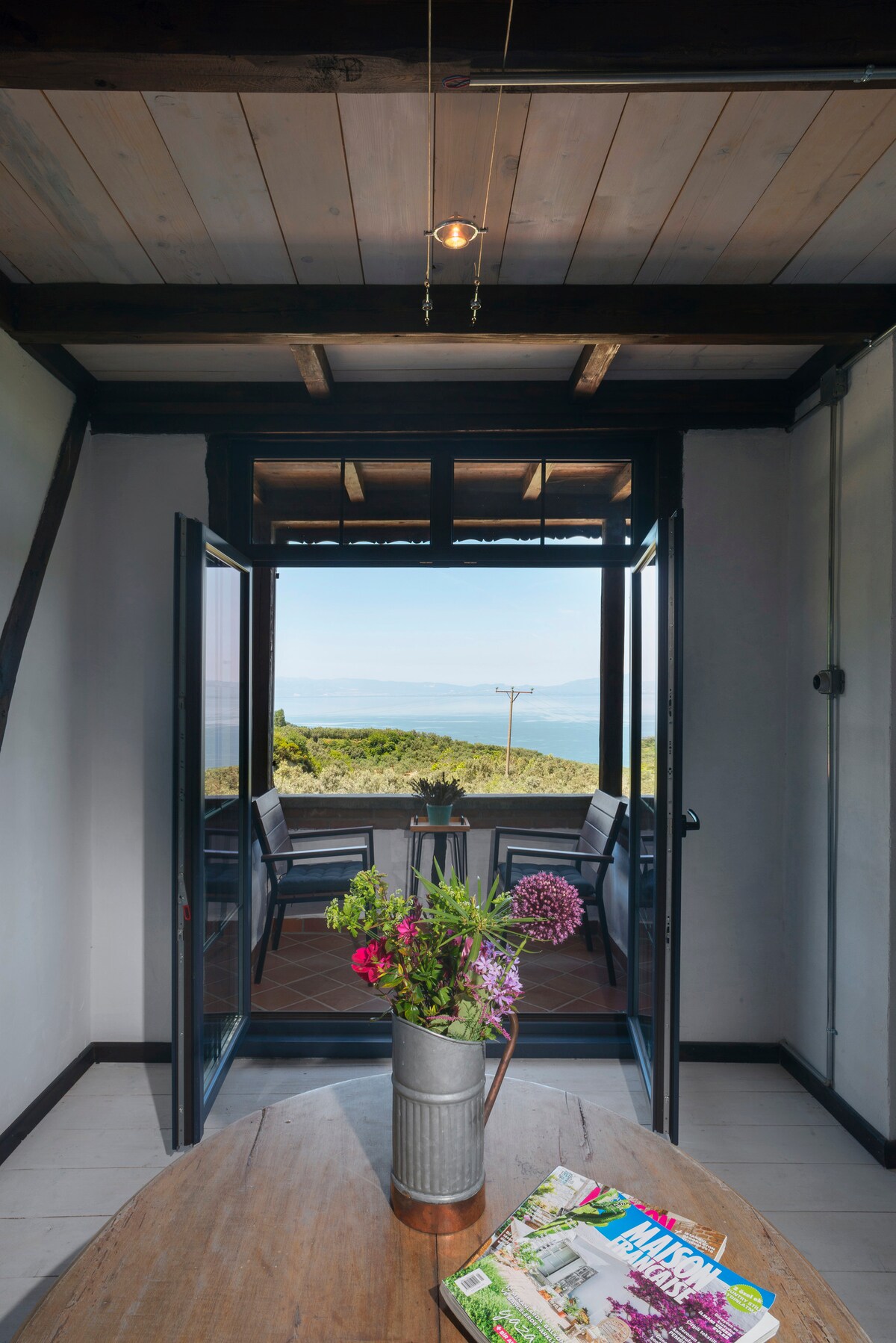 An open doorway reveals a scenic view of Lake İznik, framed by a rustic wooden frame. A round wooden table sits in the foreground, adorned with a vase of fresh flowers. Outdoor seating can be seen just beyond the threshold, enhancing the connection to nature.