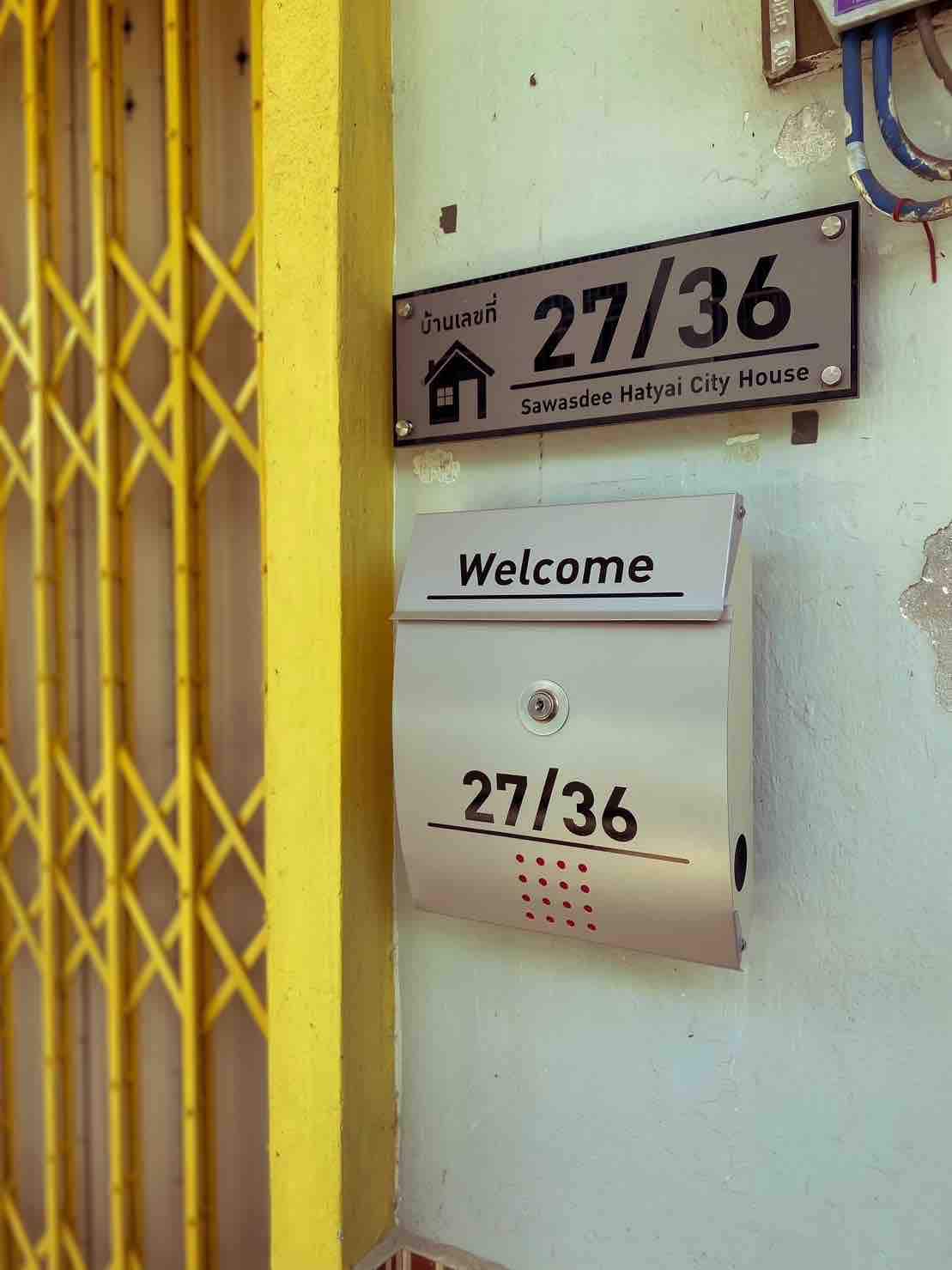 A clear and inviting entrance displays the house number 27/36, prominently featured on a modern mailbox. A welcoming sign invites guests, positioned next to a yellow security gate that adds a touch of color to the exterior.
