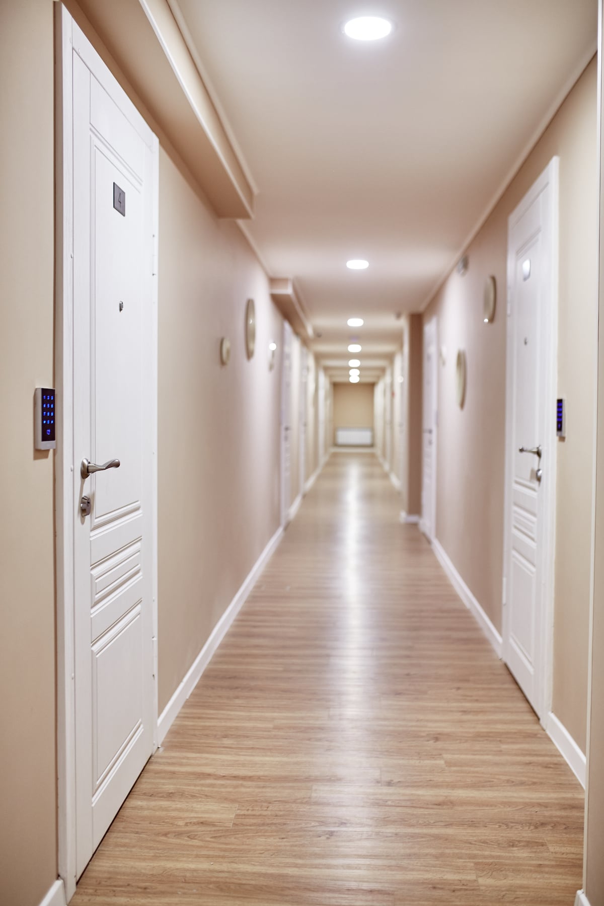 A well-lit hallway features smooth wooden flooring and light-colored walls. Doors lined along either side provide access to guest accommodations. Soft overhead lighting enhances visibility, while wall-mounted features are present for security access.