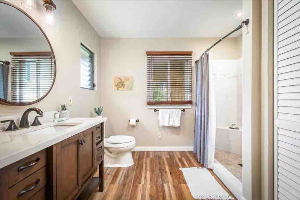 The bathroom features a large vanity with dual sinks beneath a round mirror. Natural light filters through window treatments. A walk-in shower with glass doors is visible. Wooden flooring complements the space, which includes a toilet and a small decorative plant on the counter.