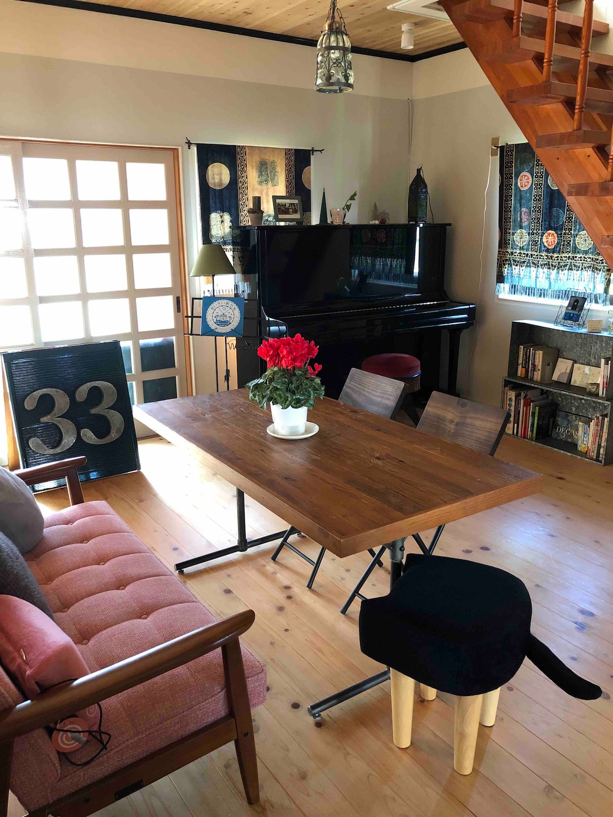 The living area displays a wooden table surrounded by chairs, accompanied by a couch in warm tones. A piano stands in the corner, while decorative curtains frame the windows. A potted plant on the table adds a touch of greenery to the space.