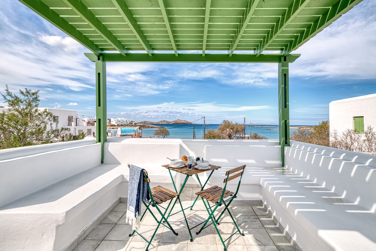 A spacious terrace is featured, framed by green beams, offering views of the turquoise waters. A small table is set with breakfast items and two wooden chairs. Built-in seating runs along the perimeter, creating an inviting space for dining and relaxation.
