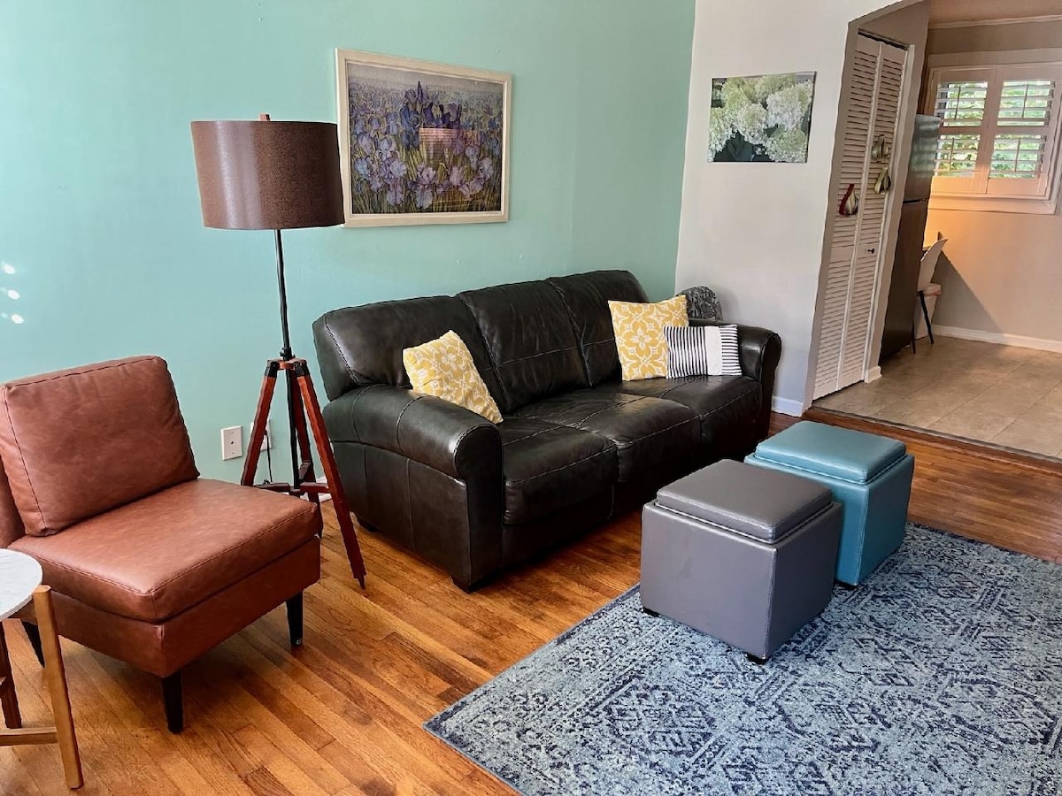 A living area features a dark leather sofa with decorative pillows alongside a mid-century modern armchair. Two ottomans in contrasting colors complement a large area rug on hardwood flooring. A painting adorns the wall, while natural light filters through partially open shutters.