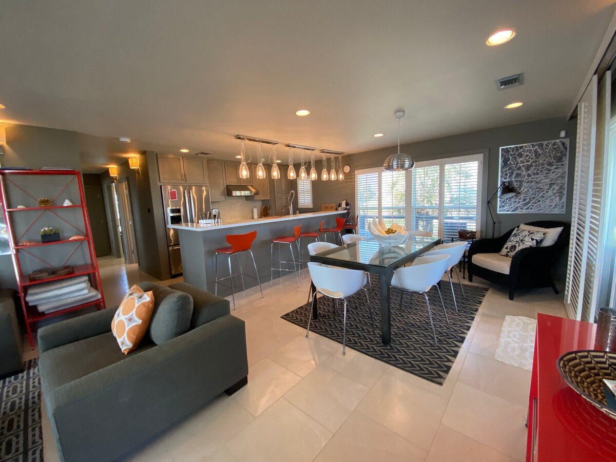 A modern living space features an open layout with a glass dining table surrounded by white chairs and colorful bar stools at the kitchen counter. A black sofa complements the decor, while large windows let in natural light, enhancing the inviting atmosphere.