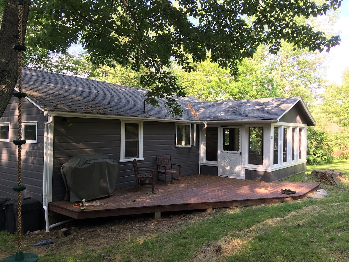 The exterior of the property reveals a charming rustic cabin with a well-maintained gray facade. A spacious wooden deck extends from the house, flanked by large windows that invite natural light. A grill is partially visible on the deck, surrounded by greenery.