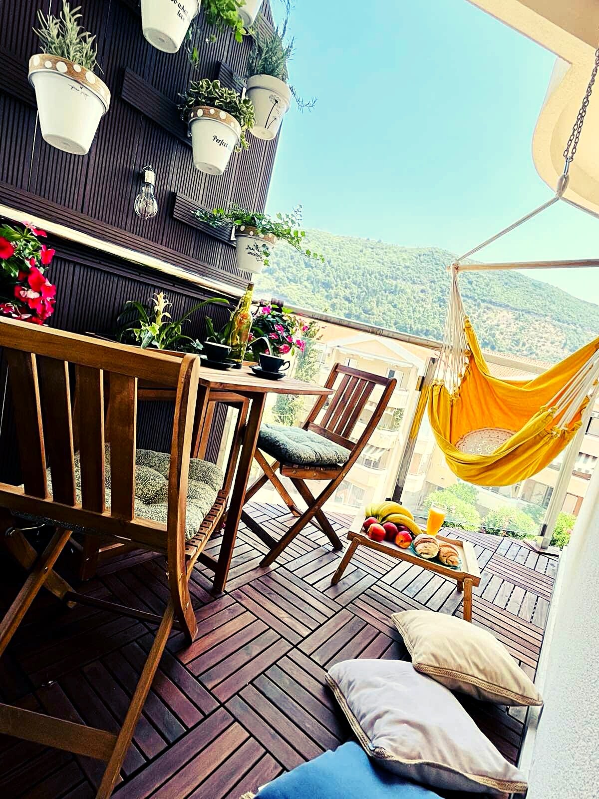 A small terrace is adorned with hanging flower pots and vibrant greenery. A wooden table with two chairs is complemented by cushions on the floor. A yellow hammock adds a relaxing touch, while a platter of fruits and a beverage sit invitingly on the table.
