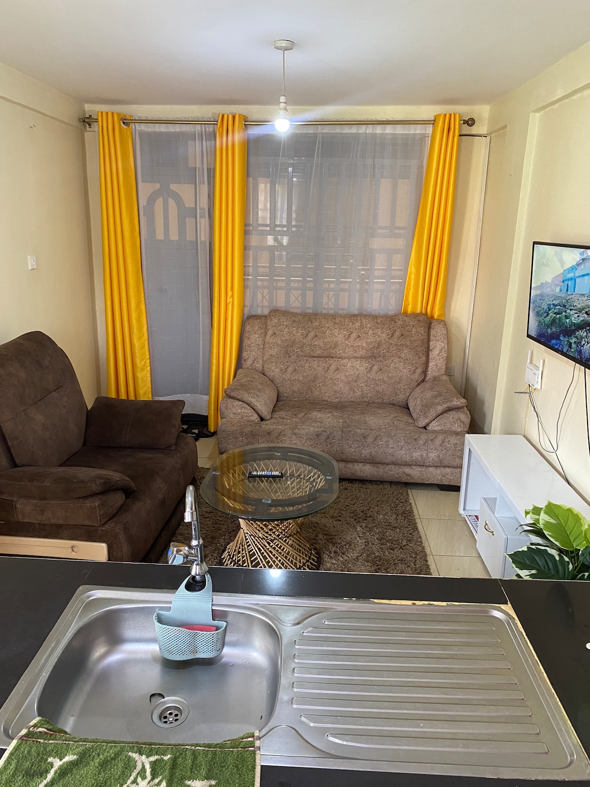 A comfortable living area features a brown couch and matching armchair beside a glass coffee table. Bright yellow curtains frame a window, allowing light to filter through. A kitchen sink and towel are visible in the foreground, providing easy access to cooking essentials.