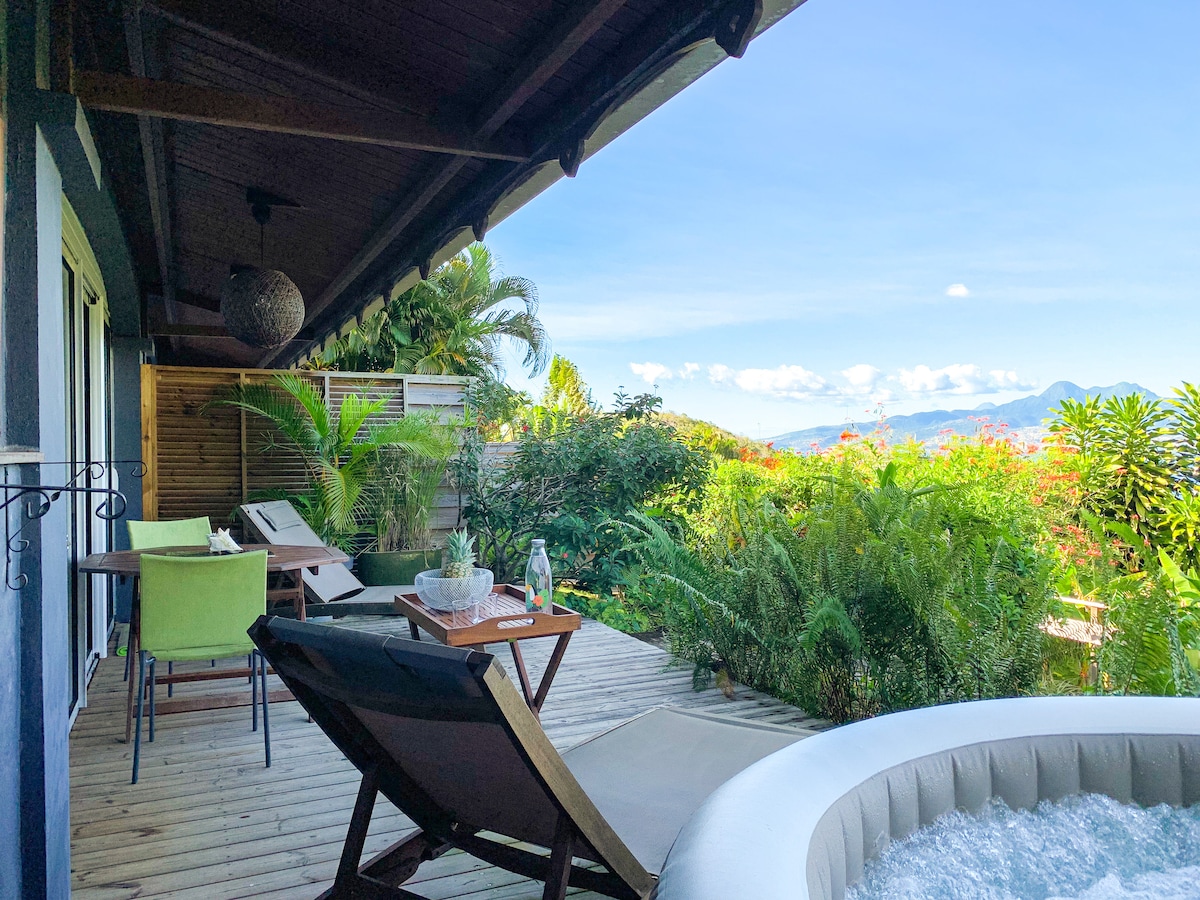 A spacious terrace is visible, featuring a lounging chair alongside a table set with a bottle and two glasses. Lush greenery surrounds the area, and a private jacuzzi is positioned in the corner, with distant mountains under a clear blue sky.