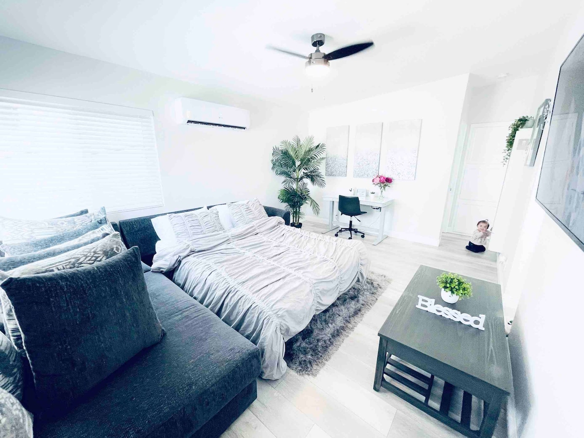 A cozy sleeping area features a spacious bed covered with light bedding, accented by plush pillows. A modern sofa is located nearby, along with a desk featuring a chair and a small plant. Natural light fills the room through a window with white blinds.