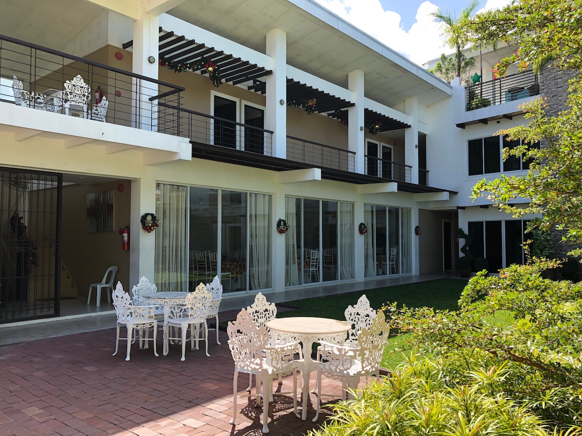 An outdoor seating area features elegant white furniture arranged around a circular table. The space is bordered by a lush green lawn and is framed by a two-story building with large glass doors and shaded balconies. Natural light enhances the inviting environment.