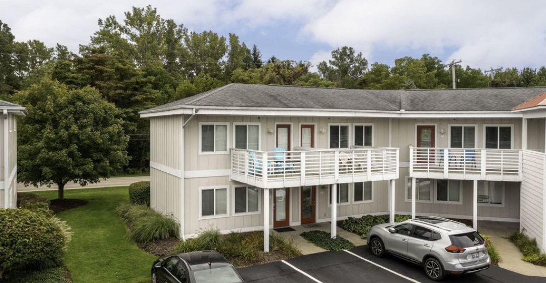 A two-level duplex condo is shown amidst lush greenery. The exterior features multiple windows and two inviting balconies. A designated parking area is visible in the foreground, along with well-maintained landscaping and trees around the property.