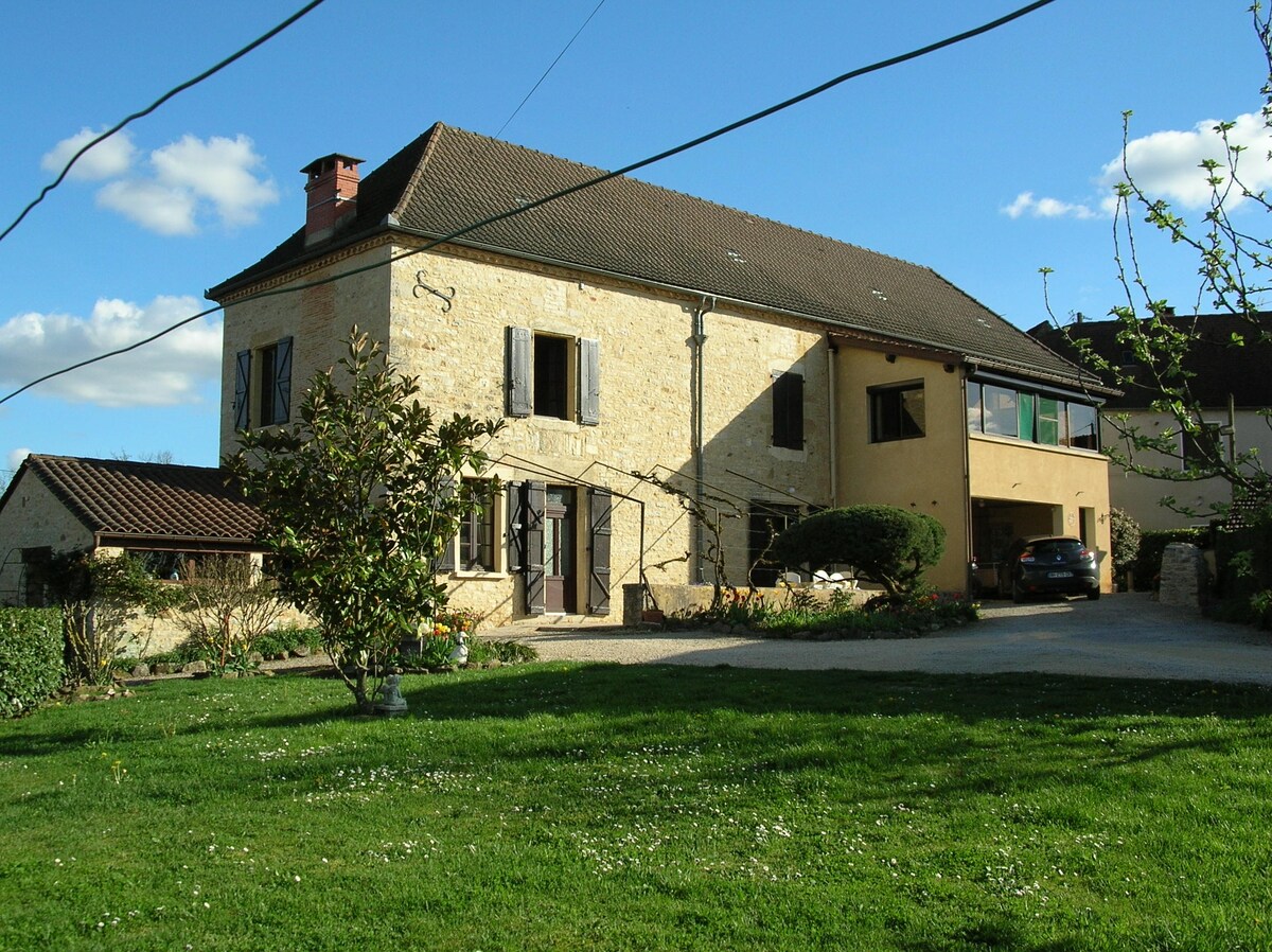 An elegant stone house is framed by a large green lawn, featuring symmetrical windows and a combination of shutters. A spacious driveway is visible, along with a vehicle parked nearby. The structure boasts multiple stories, with a well-maintained garden accentuating the welcoming exterior.