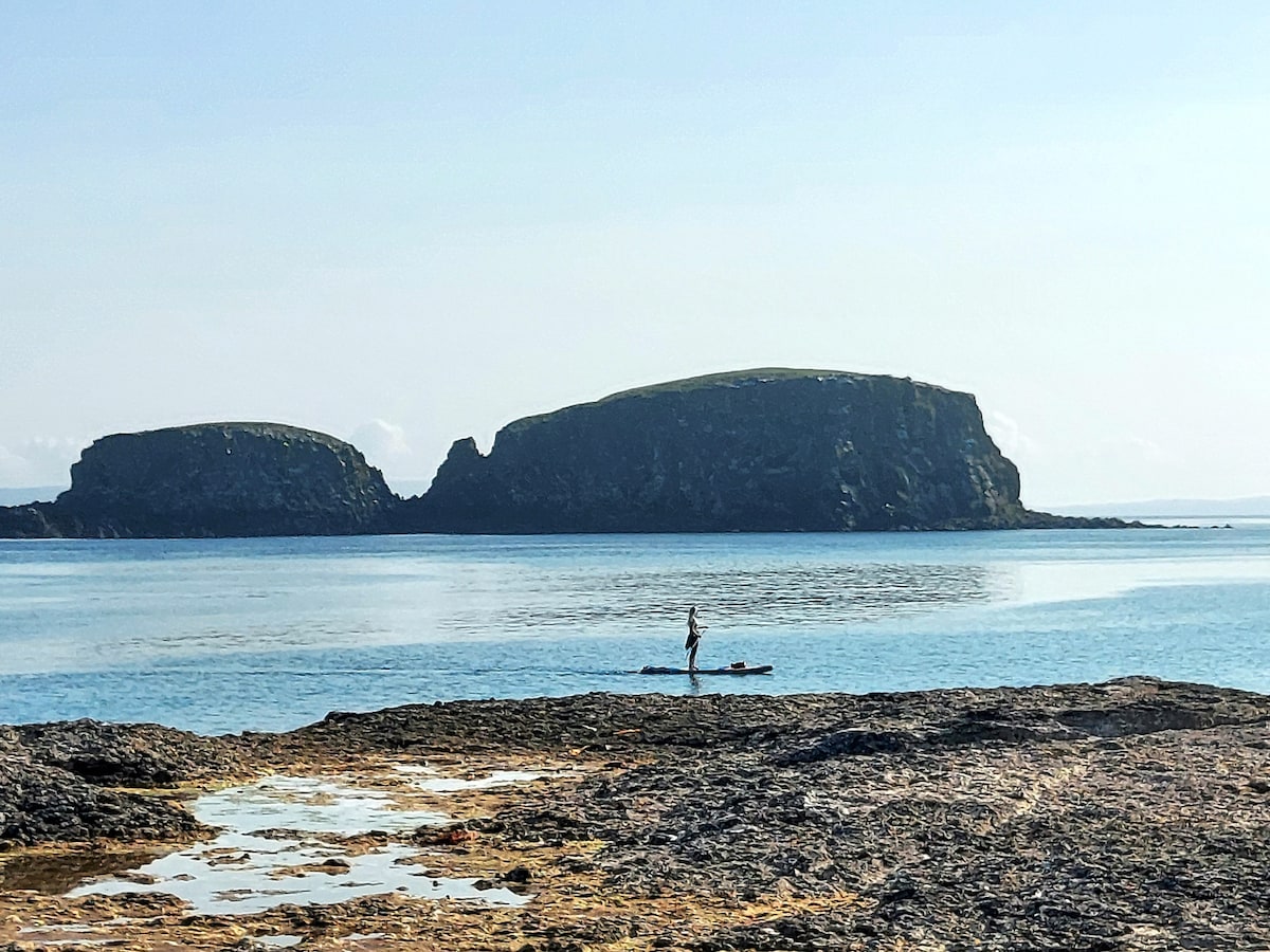 A tranquil coastal scene is presented, showcasing calm blue waters with a person kayaking in the foreground. Distinctive rocky formations rise in the background, surrounded by shimmering sea. The sky is clear, enhancing the serene ambiance of the waterfront landscape.