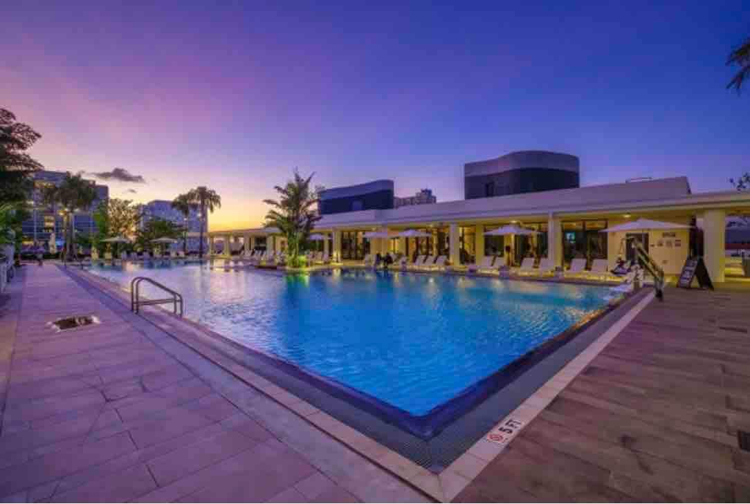 The infinity pool glistens with vibrant blue water, surrounded by lounge chairs and palm trees. A serene atmosphere is created by the soft lighting and the twilight sky, which complements the modern architecture of the nearby building.