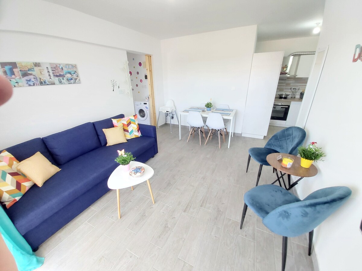 A bright living area features a blue sofa adorned with colorful cushions, accompanied by a round coffee table. A dining table with four white chairs is positioned nearby, while the kitchen area is visible in the background, showcasing modern appliances.
