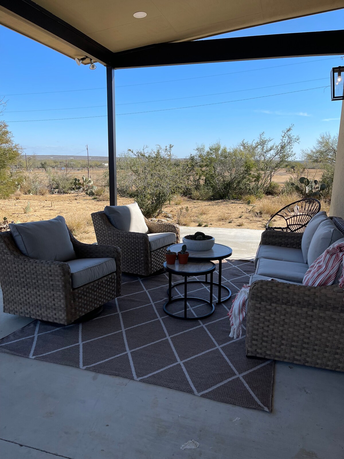 A covered outdoor seating area features plush chairs and a small round table arranged on a geometric-patterned rug. The surrounding landscape includes low vegetation and open spaces, with clear blue skies above.