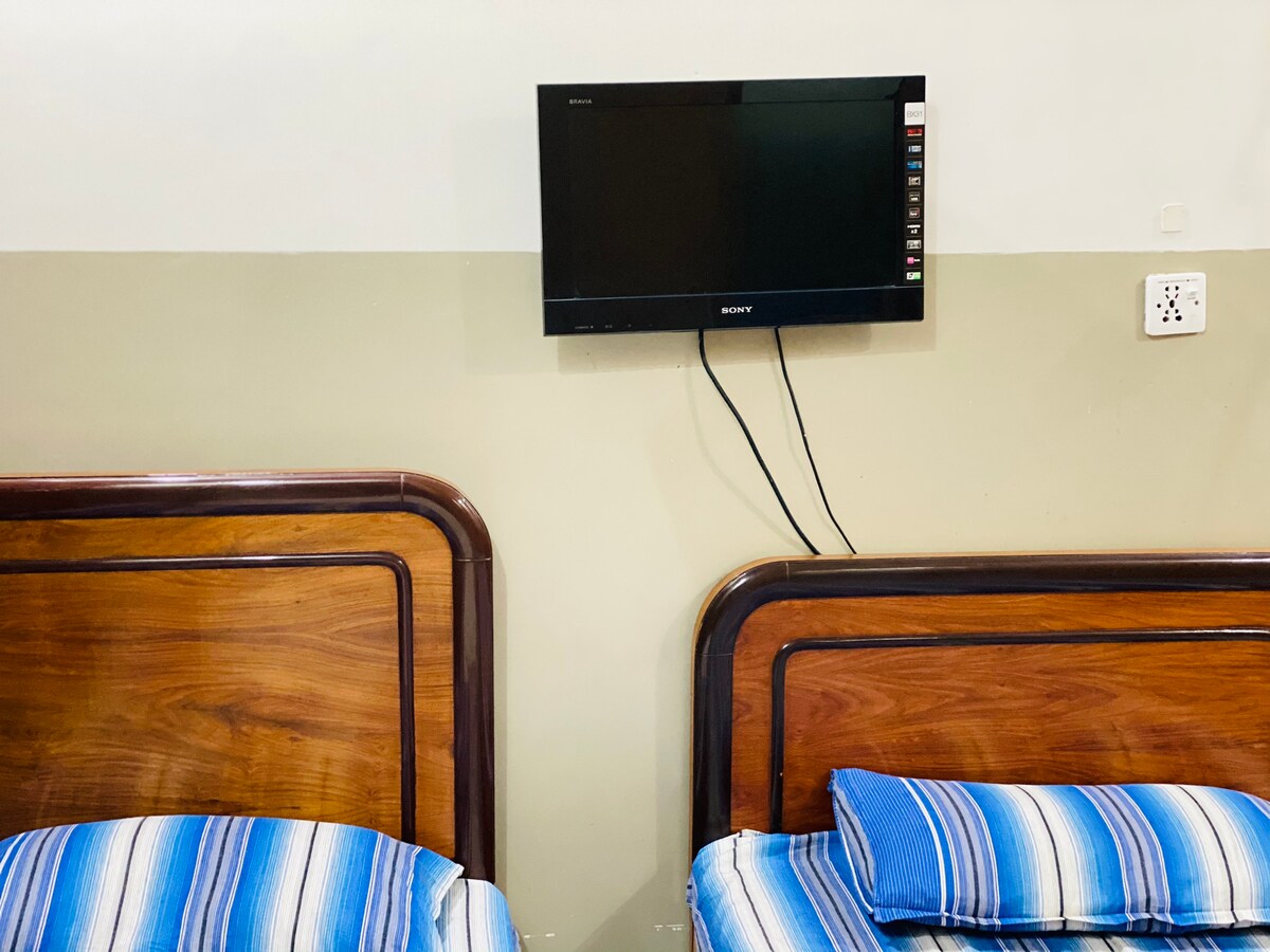 Two beds are positioned side by side, each adorned with blue striped bedding. A flat-screen television is mounted on the wall above, providing entertainment options. The warm wooden bed frames contrast with the neutral-colored wall behind them.
