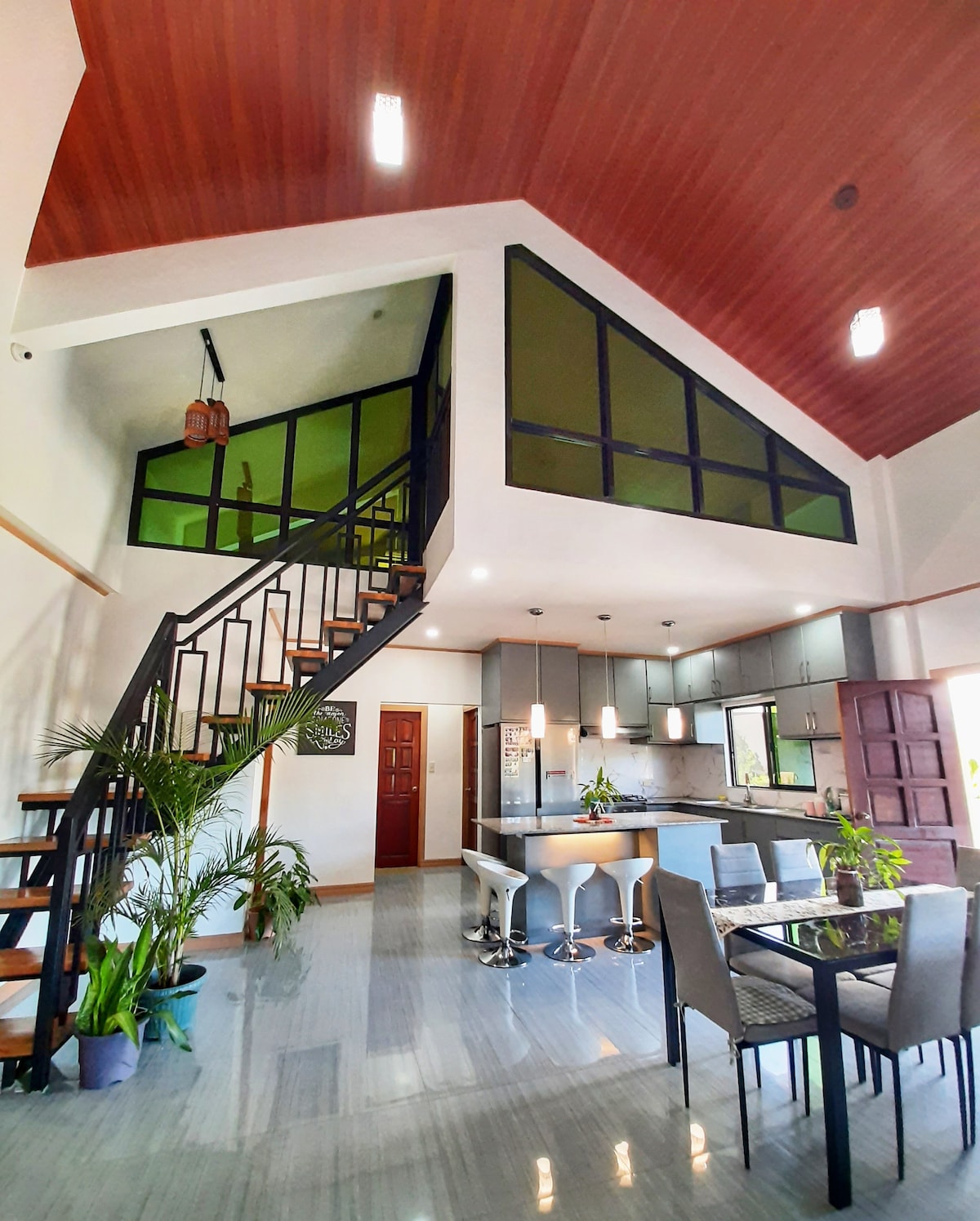 An open and airy living space features high ceilings and large windows, allowing natural light to fill the room. A modern kitchen with sleek countertops is visible, alongside a dining area equipped with a table and chairs. A staircase leads to a lofted area, enhancing the overall spaciousness.