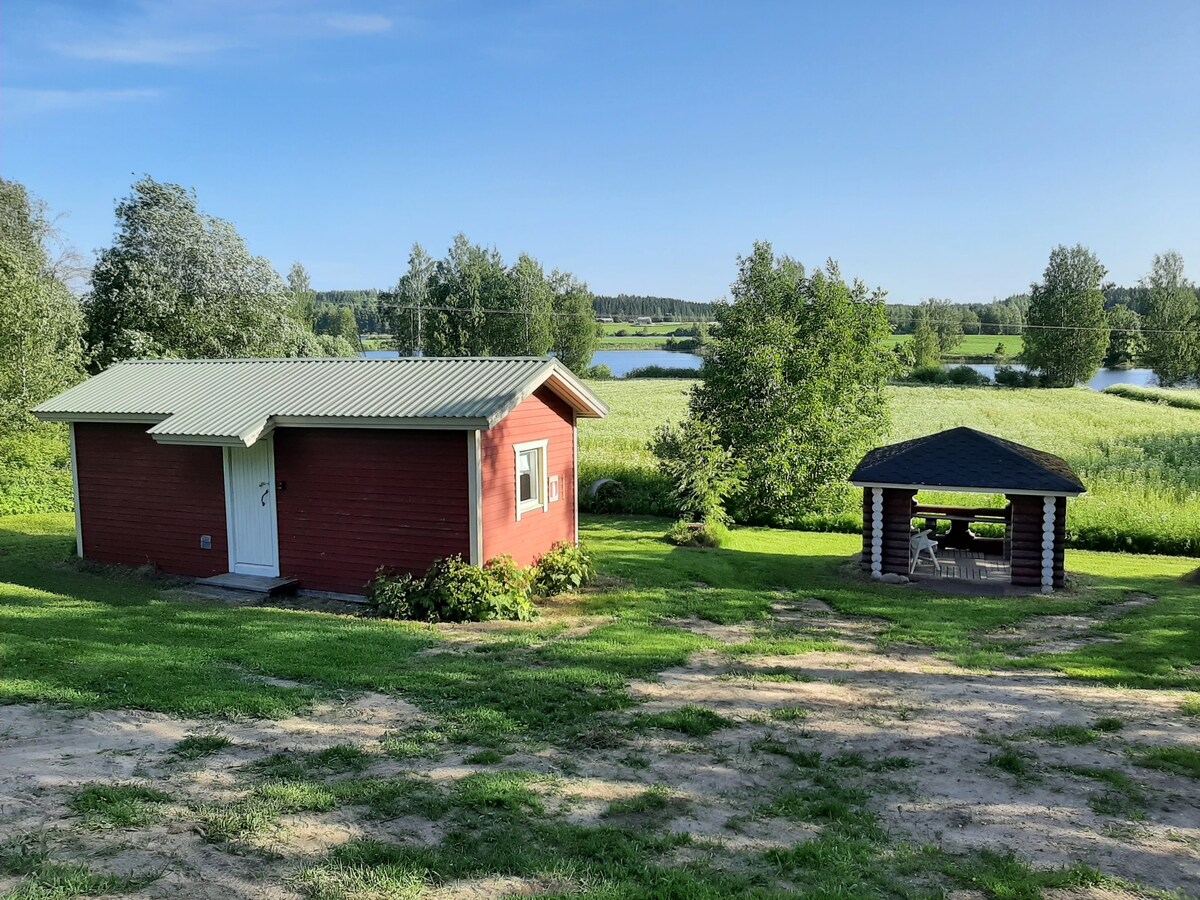 A red wooden cabin is set against a backdrop of lush green grass and trees, with a serene lake visible in the distance. Nearby, a gazebo with seating creates an inviting outdoor space for relaxation or gatherings.
