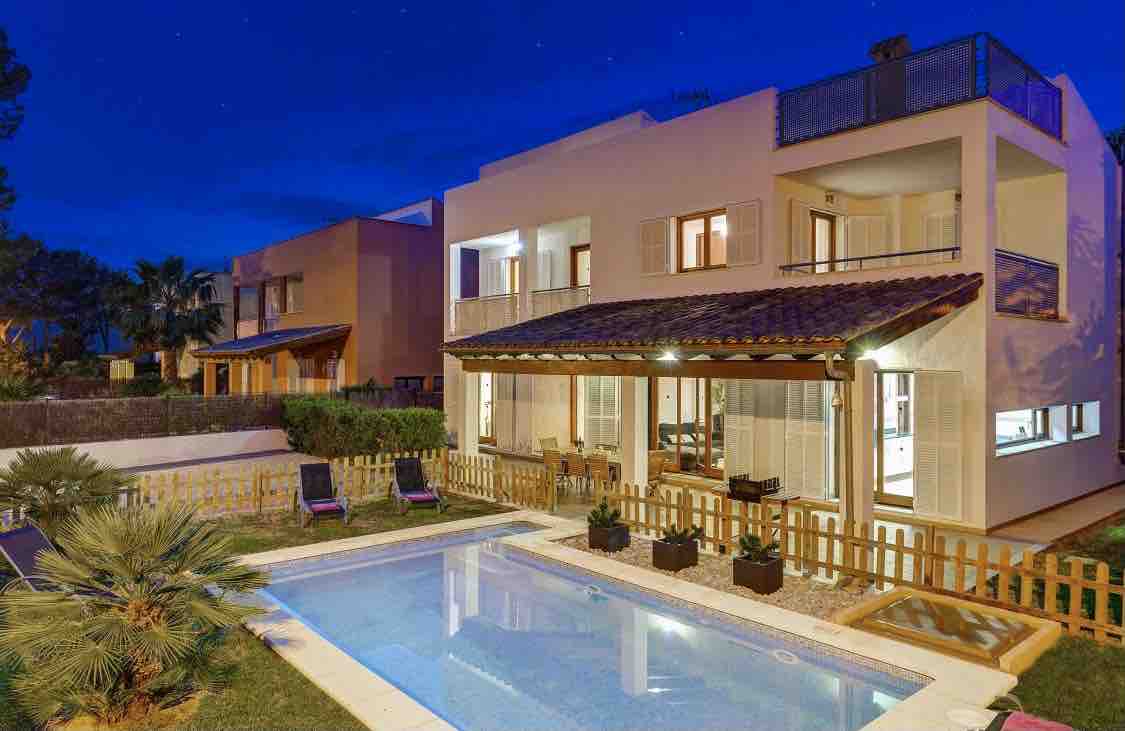 The villa features a modern exterior illuminated at dusk, showcasing large windows and a spacious terrace. A fenced pool area is visible, surrounded by potted plants and lounge chairs, set against a backdrop of lush greenery.