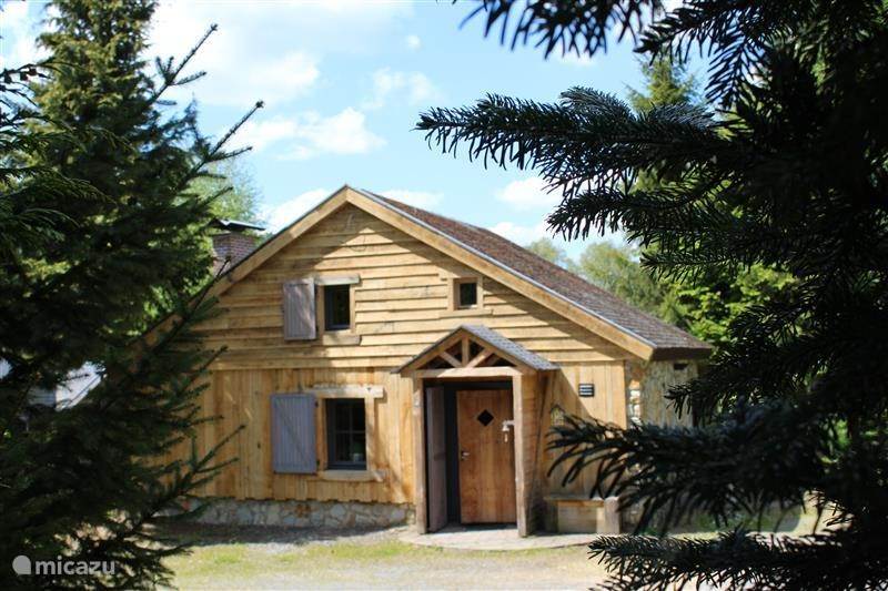 The wooden chalet is framed by lush evergreen trees, showcasing a welcoming entrance with a diamond-shaped window above the door. The façade features natural wood panels and shutters, blending harmoniously with the surroundings.