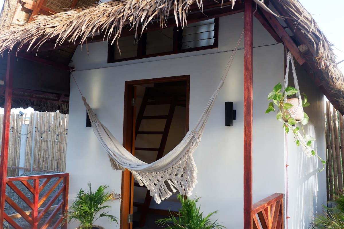 A welcoming entrance showcases a charming hammock suspended from the porch of a modern kubo. The structure features natural wood accents and is surrounded by greenery, with a rustic thatched roof enhancing its beachside appeal.