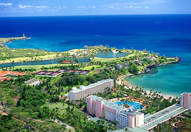 An aerial view showcases the vibrant blue ocean meeting the coastline, with lush greenery and scattered buildings along the shore. A serene lagoon is visible, surrounded by manicured landscapes. The scene reflects a harmonious blend of nature and resort amenities.
