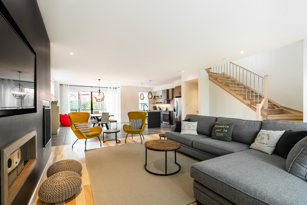 An open-plan living area is visible, featuring a gray sectional sofa paired with a round coffee table. Yellow chairs add a pop of color, with a modern TV mounted on a black wall. A staircase leads to the upper level, enhancing the bright and airy atmosphere.