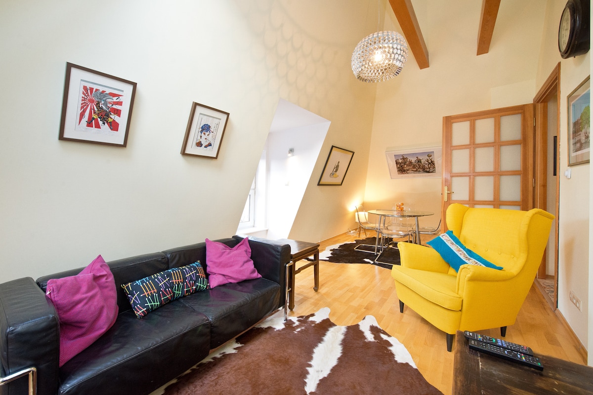 A well-furnished living area features a sleek black sofa adorned with vibrant pink cushions. A bold yellow armchair provides a pop of color. Natural light filters through large windows, illuminating the space, while decorative art pieces adorn the walls, complementing the wooden beams above.