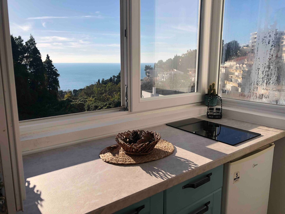 A modern kitchen area features a countertop with a cooking surface and a woven basket displayed. Large windows provide a view of the ocean and surrounding greenery, allowing natural light to illuminate the space.