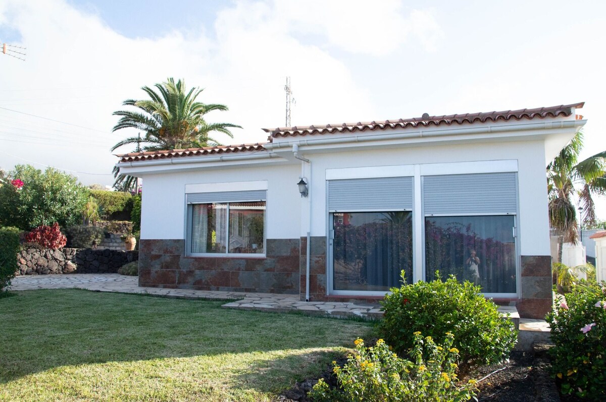 A one-story house is set within a well-kept garden, featuring lush greenery and vibrant flowers. Large windows are framed with shutters, allowing natural light to brighten the interior. A stone pathway leads to the entrance, surrounded by neatly trimmed grass.
