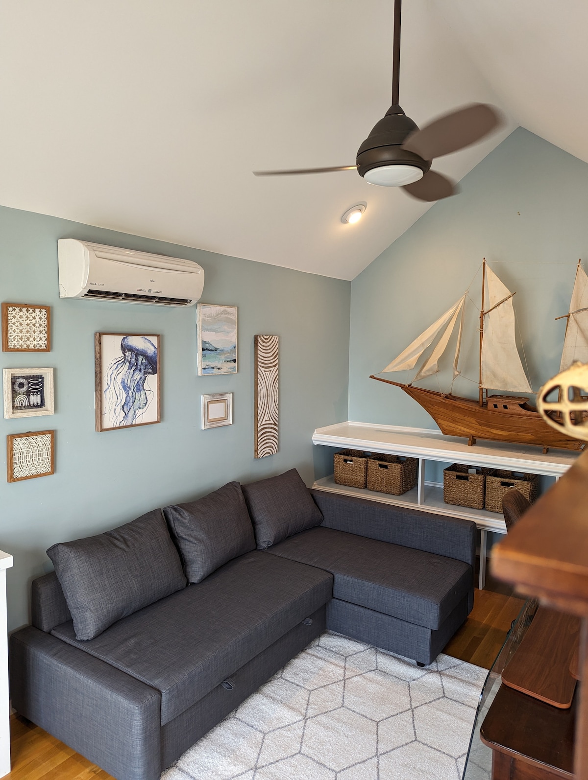 A cozy corner of the room features a gray sectional couch complemented by a patterned area rug. The light blue walls are adorned with framed artwork and nautical decor, including a model sailboat displayed on a shelf. A ceiling fan is positioned above for comfort.