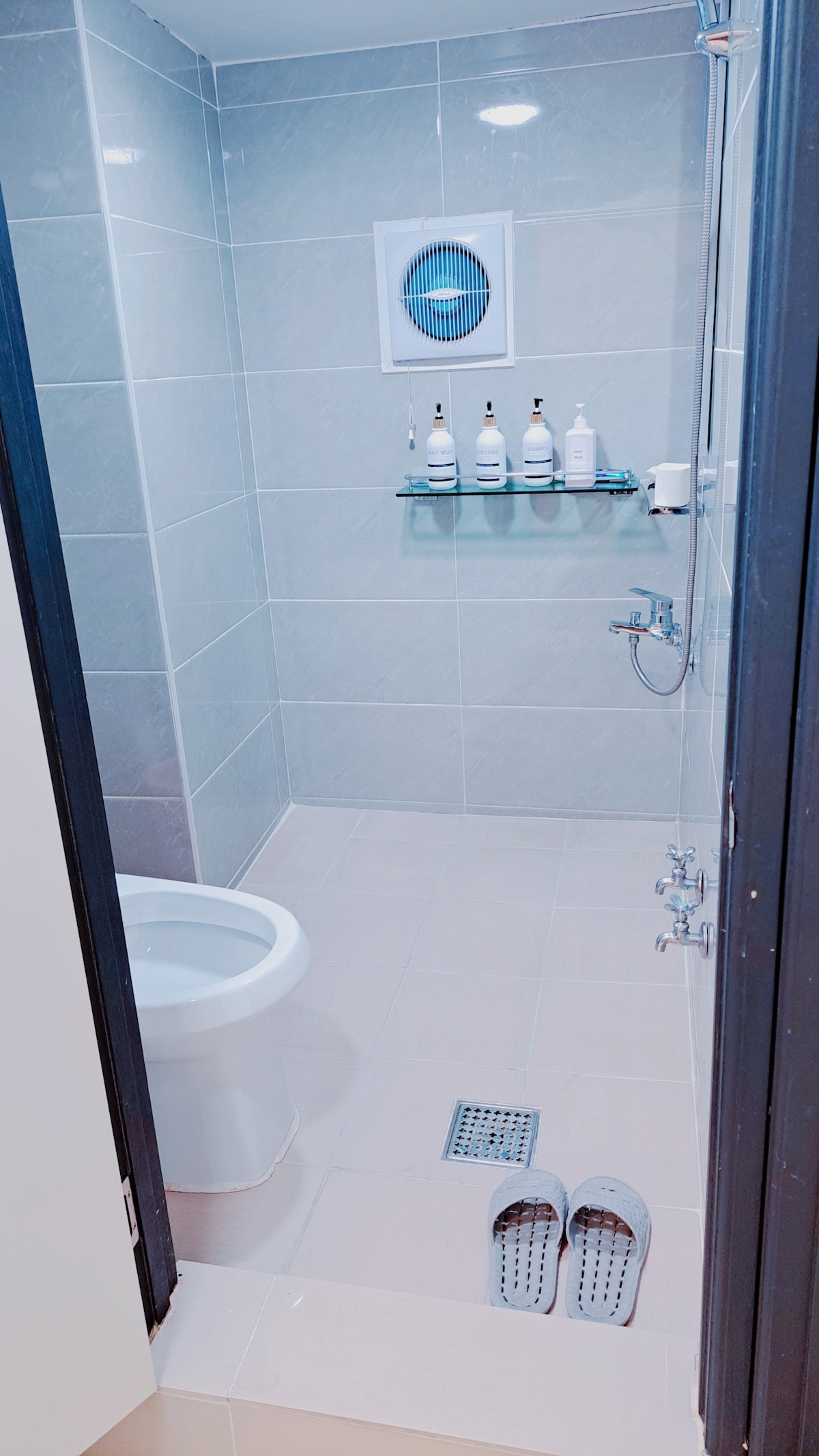 A bright and clean bathroom is displayed, featuring a modern toilet and a walk-in shower with transparent glass. A small green shelf holds several bottles of toiletries, and a drain is visible on the light-colored floor. A pair of slippers is placed neatly at the entrance.