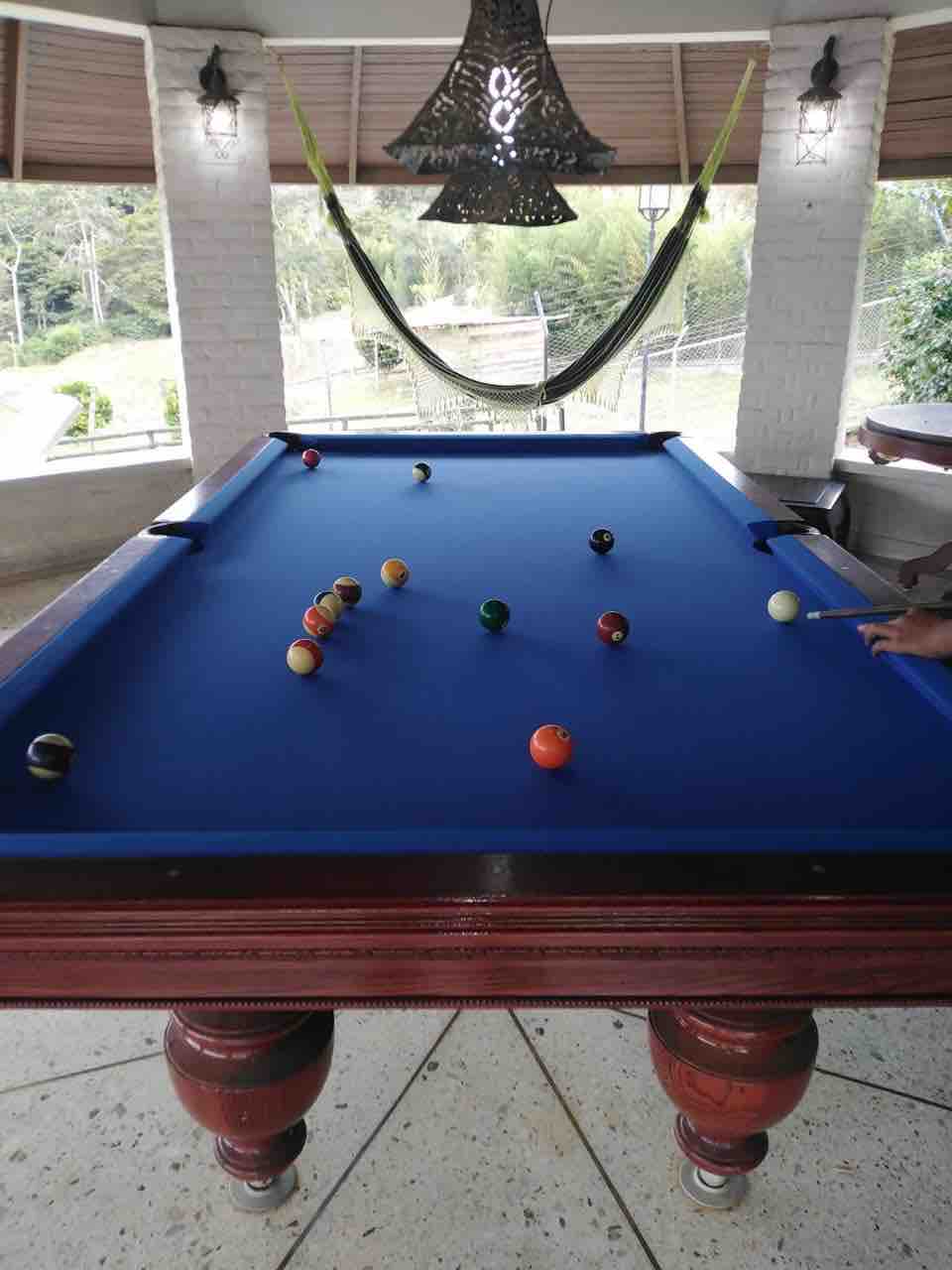 A billiards table is situated in a shaded area, featuring a vibrant blue felt surface and polished wooden legs. Several colorful pool balls are arranged on the table, with a cue stick poised for play. A hammock is visible in the background, adding to the relaxed atmosphere.
