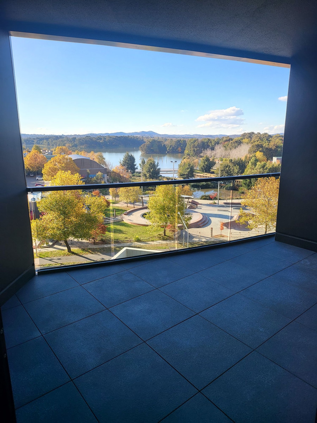 An expansive balcony offers a scenic view of a lake surrounded by autumnal trees. The foreground features well-maintained pathways and landscaped areas, enhancing the tranquil environment. Clear skies and gentle sunlight set a peaceful backdrop.