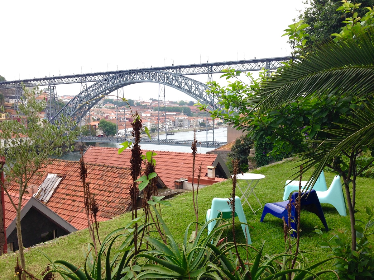 A lush garden area is visible, featuring green grass and a variety of plants. A small table and colorful chairs are placed on the lawn, offering a view of the iconic bridge over the Douro River, surrounded by terracotta-roofed buildings.
