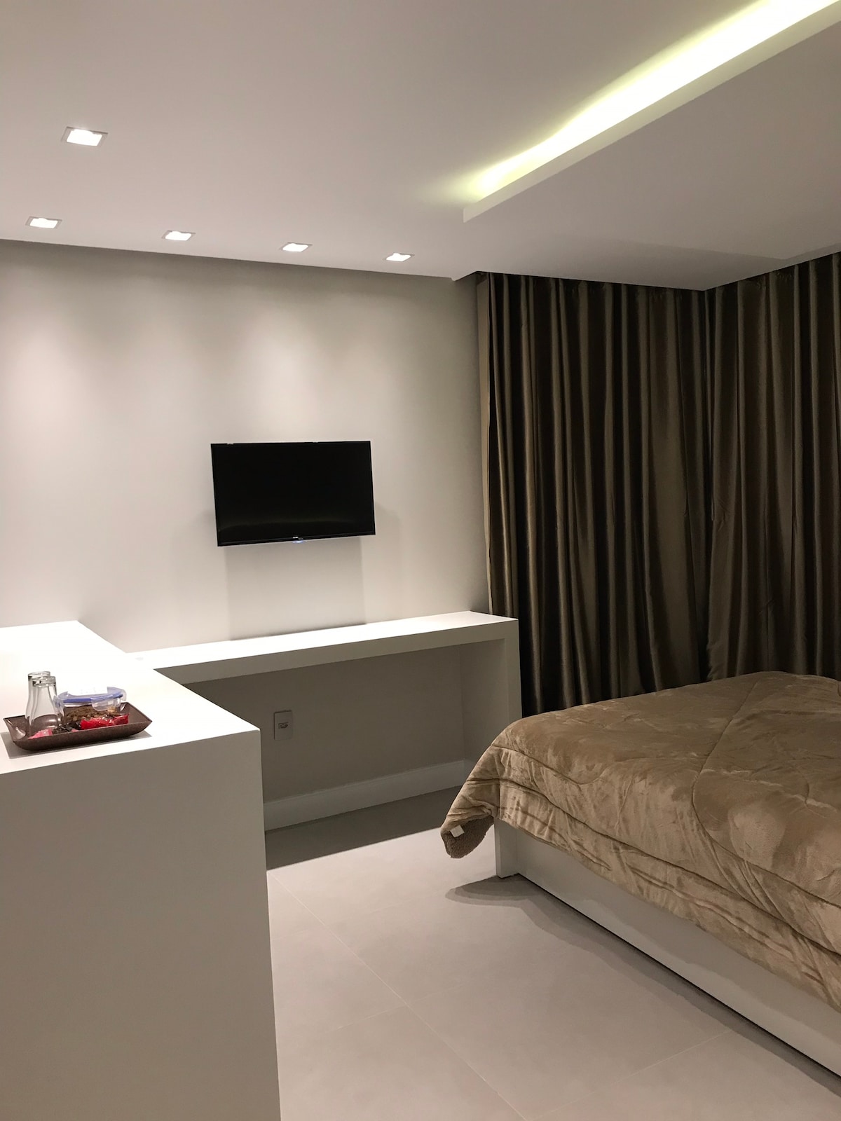 A well-lit bedroom features a spacious bed covered with a soft beige quilt. A small white desk is positioned opposite a wall-mounted television. Floor-to-ceiling curtains in a deep hue frame the window, contributing to the modern and minimalistic design.