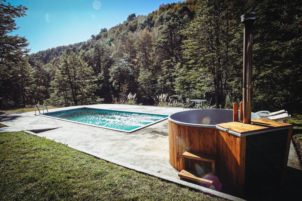 A tranquil outdoor area features a swimming pool with clear water alongside a wooden hot tub, showcasing their natural surroundings. Lush green grass and trees provide a serene backdrop, while a patio area with steps leads to the hot tub, inviting relaxation.