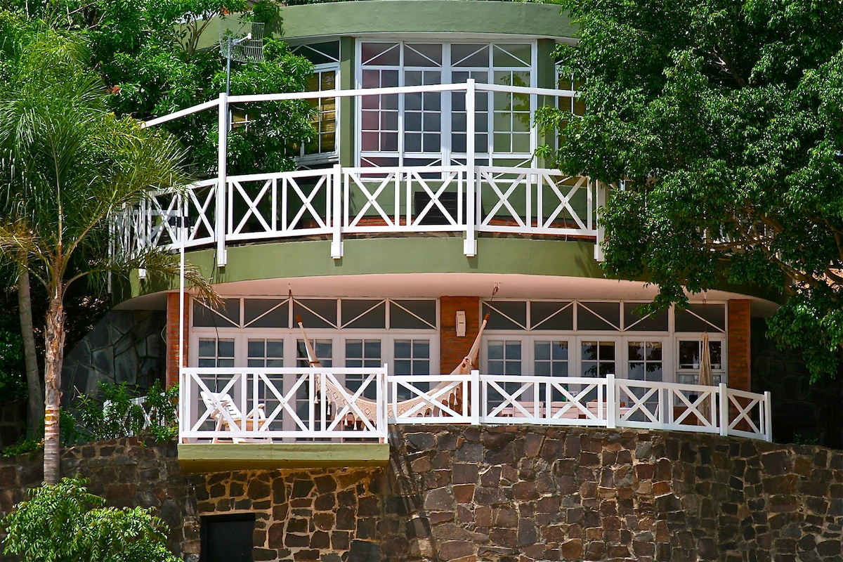 The property features a rounded architecture with large glass windows enclosed by a white railing. A spacious deck is visible, bordered by lush greenery and a stone foundation, providing a connection to the surrounding landscape.