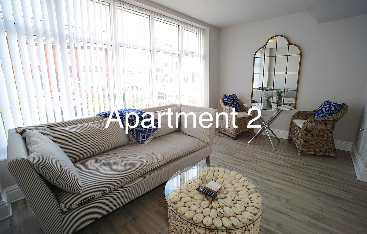 A bright lounge area is featured, with large windows allowing ample natural light. A comfortable couch is complemented by two wicker chairs, each adorned with decorative blue cushions. A round coffee table, decorated with shells, sits in the center of the space.