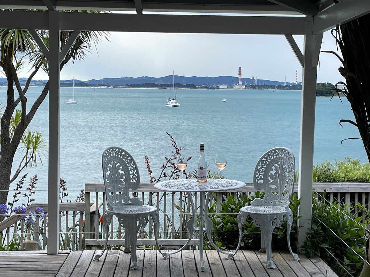 A small outdoor seating area features a round table with two white, intricately designed chairs. A bottle and glasses sit atop the table, with a scenic view of the bay and distant boats visible beyond. Lush greenery frames the space, enhancing the tranquil atmosphere.