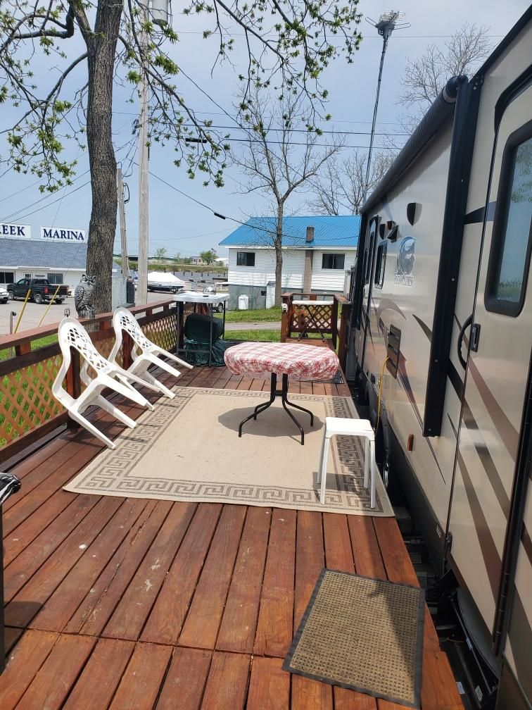 A wooden deck is adorned with a round table and two white chairs. An area rug adds texture, while surrounding trees provide shade. The nearby marina is visible in the background, creating a relaxed outdoor space ideal for gathering.