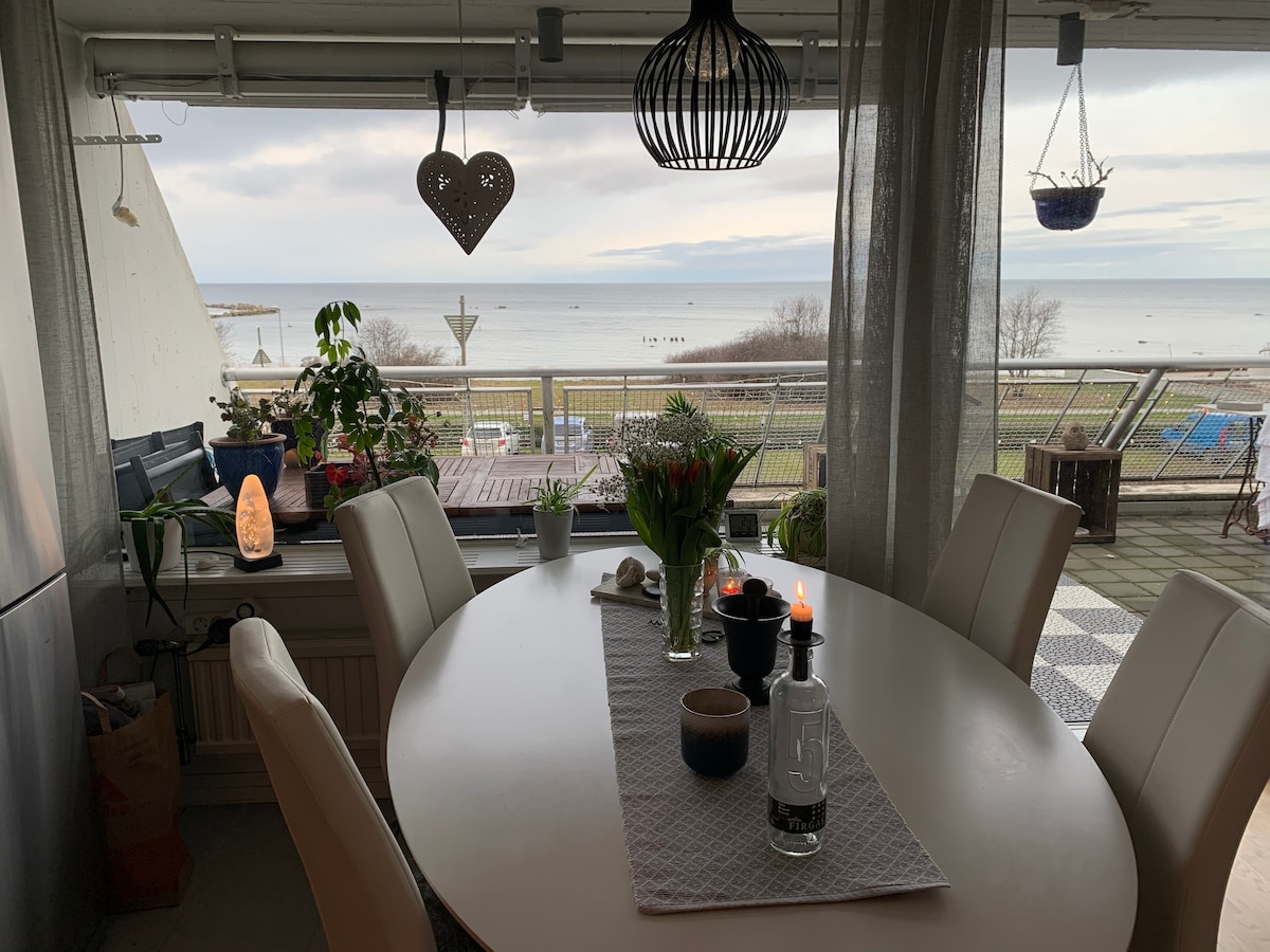 A dining area features a round table surrounded by white chairs, with a view of the sea visible through large windows. Potted plants and candles adorn the table, enhancing a calm atmosphere. The outdoor area displays greenery and the water in the distance.